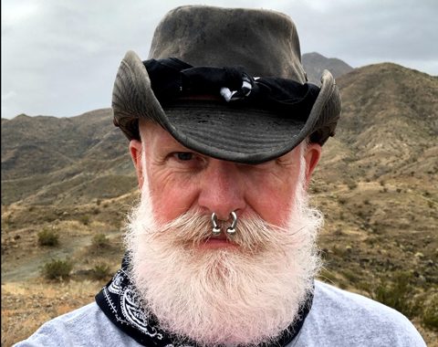 A rugged man with a white beard, black hat, and desert landscape, highlightin
