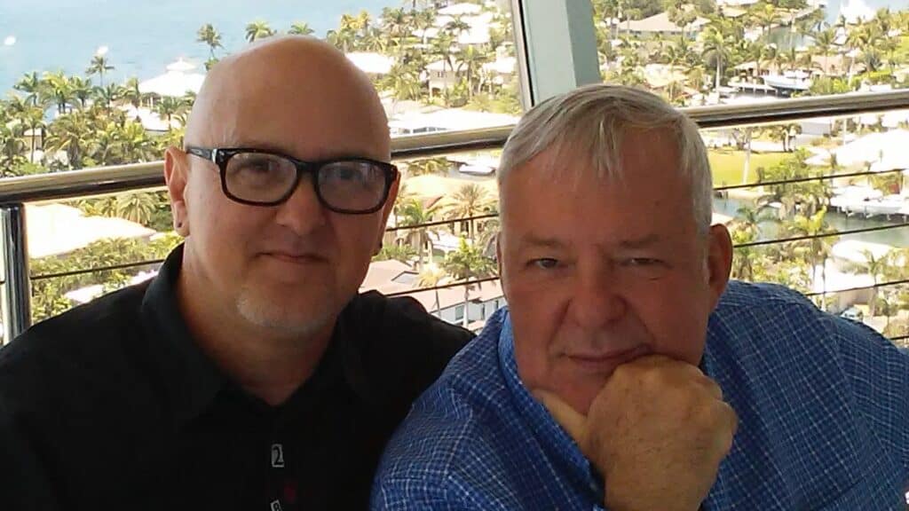 Bald man with glasses and another man in blue shirt sitting outdoors overlooking a tropical resort.