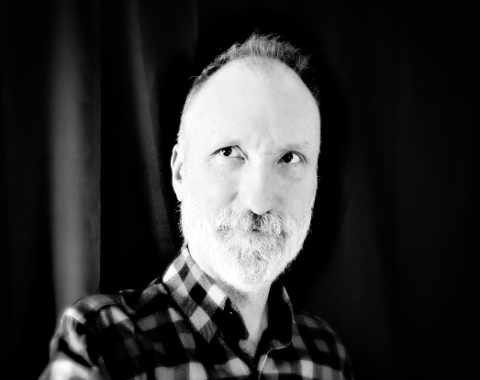Black and white portrait of a man with a beard and checkered shirt, promoting Rattling Good Yarns Press.