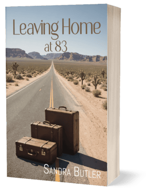 The front cover of "Leaving Home at 83" by Sandra Butler. The cover depicts a long road through the desert disappearing into the horizon. In the middle of the road are three vintage-style suitcases.