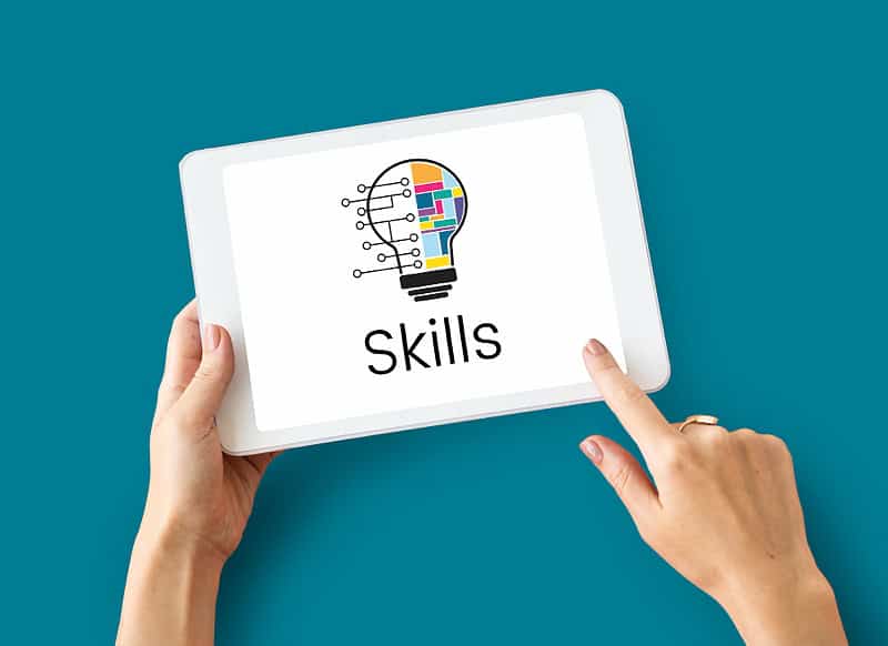 A person holding a tablet with a graphic of a light bulb made of digital circuit lines and colorful puzzle pieces, above the word "Skills" on the screen. The background is solid teal.