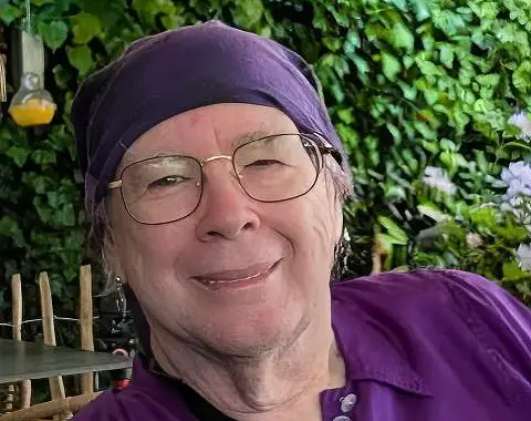 A close-up photo of author Jenny Yates, whose work explores queer love and humanity's resilience. She wears a purple headscarf and glasses, smiling against a backdrop of garden foliage. Her novel Birdless tells the story of love and hope during Earth's final disintegration.