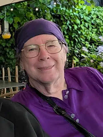 Author headshot of Jenny Yates. She wears wire-rimmed glasses, a purple headscarf, and a vibrant purple blouse, smiling warmlytoward the camera. Behind her, garden foliage and small white flowers create a soft, natural setting.