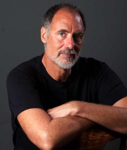 A waist-up portrait of Louis Flint Ceci, author of "The Cross Quarter Mysteries." He is a man with short salt-and-pepper hair and a groomed gray beard, wearing a black t-shirt. He is posed with his arms crossed, leaning forward against the back of a wooden chair. He looks directly at the camera with a calm, friendly expression against a dark gray background.
