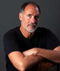 A waist-up portrait of Louis Flint Ceci, author of "The Cross Quarter Mysteries." He is a man with short salt-and-pepper hair and a groomed gray beard, wearing a black t-shirt. He is posed with his arms crossed, leaning forward against the back of a wooden chair. He looks directly at the camera with a calm, friendly expression against a dark gray background.